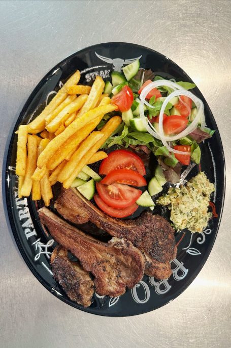 Lammkoteletts mit Pommes & Salat Ein schwarzer Teller mit Pommes, Salat, Tomaten, Gurken und gebratenem Fleisch.