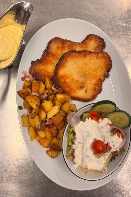 Béarnaise-Schnitzel mit Bratkartoffeln Frittiertes Fleisch mit gebratenen Kartoffeln, Salat, Gurken und einer Soße.