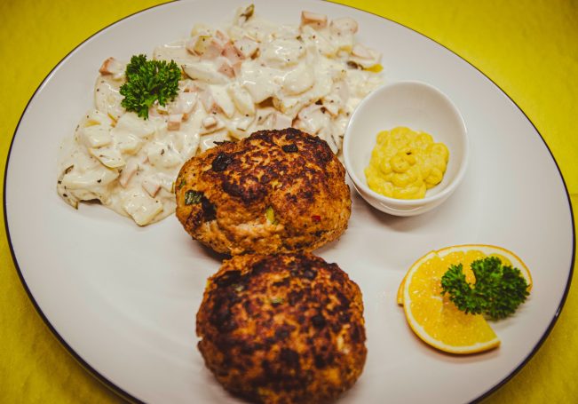 Frikadellen mit Kartoffelsalat Zwei goldbraune Fischfrikadellen mit Remoulade, Salat und Zitronenscheiben auf einem Teller.